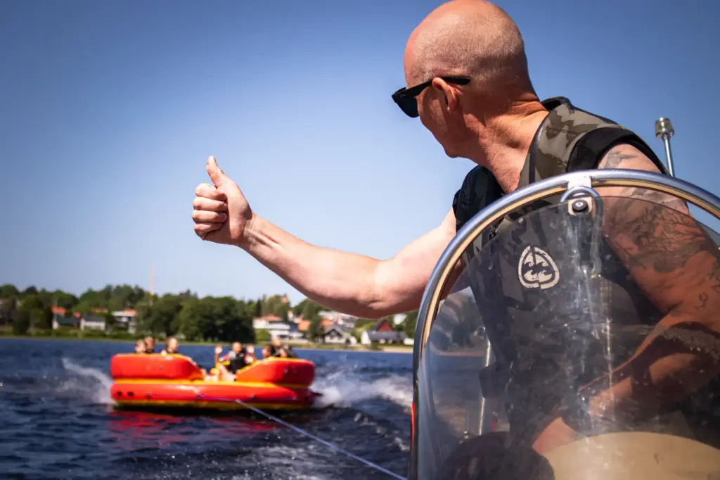 Kille kör båt och gör tummen upp åt ett gäng på en madrass efter båten.