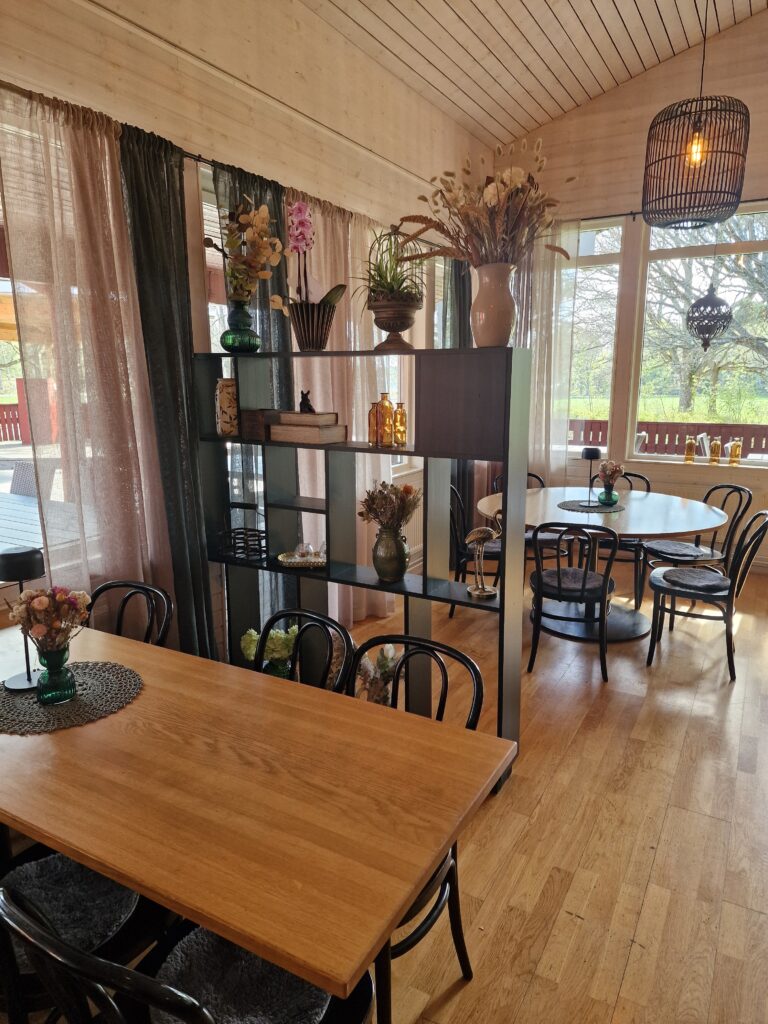 En matsal med fina träbord och mörka stolar, stora fönster med gardiner. En hylla delar av rummet lite och där står dekorationer som blommor, skålar och vaser