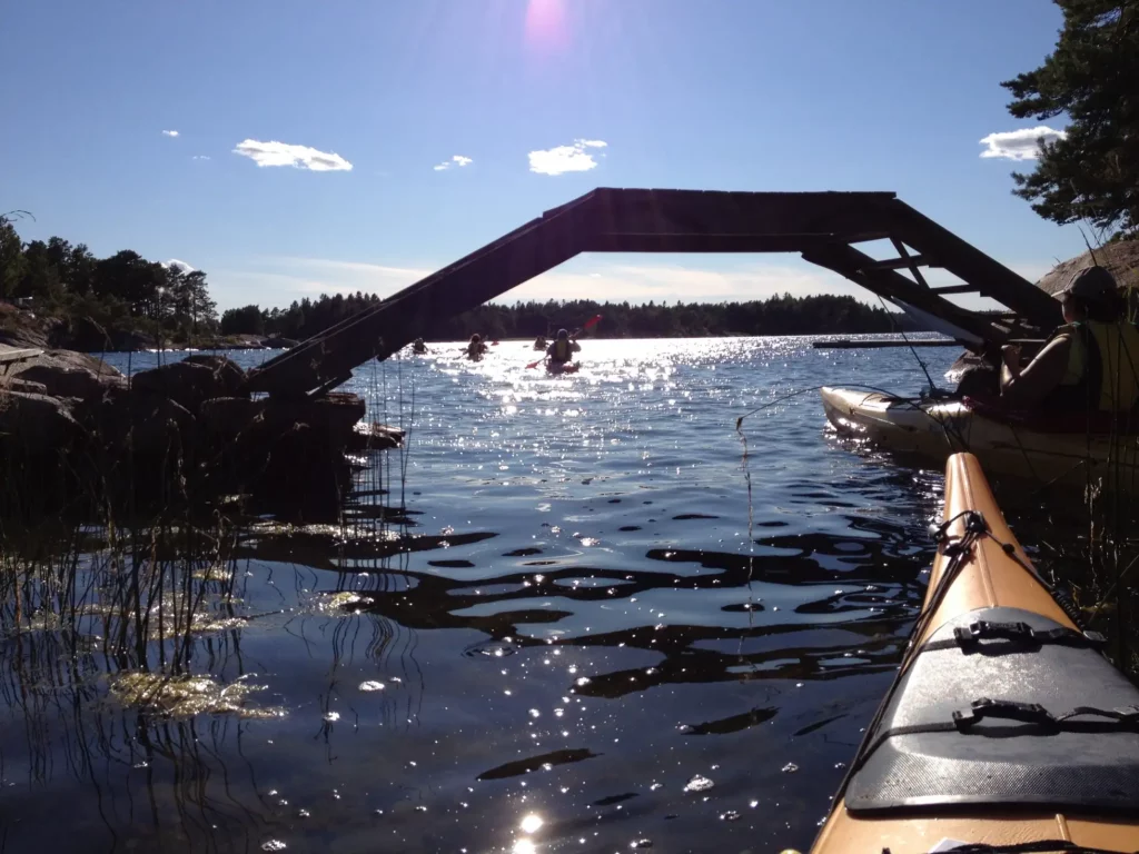 fören på en kajak syns mot ett vattendrag vid en bro. Solens ljus reflekteras mot vattnet dubbelkajaker i skärgården