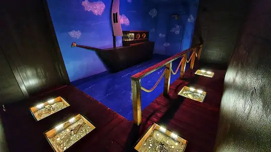 Ett rum med pirat-tema där man står på en brygga och ser ett piratskepp på avstånd. I bryggan finns rutor av plexiglas över med sand och guldmynt