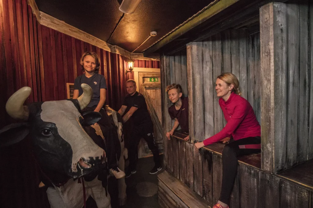 En pojke sitter på en kossa av plats och två personer tittar fram ut varsitt bås, som ser ut som dass