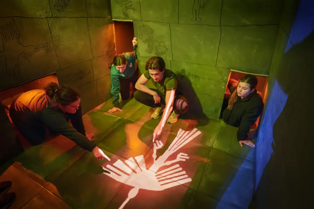 tre personer sticker fram ut varsin öppning mot et litet rum där det är en stor symbol av en fågel på golvet. En kille sitter i rummet och pekar på symbolen.