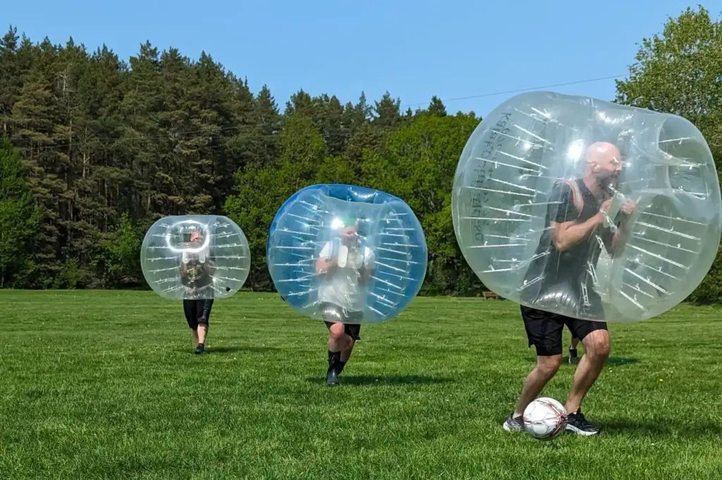 Tre män har på sig varsin stor boll springer efter en fotboll på en gräsmatta